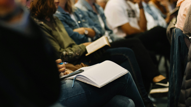 Grupa cilvēku sēž auditorijā vai seminārā, daži raksta piezīmes kladēs, citi lasa grāmatas. Priekšplānā redzams cilvēks ar atvērtu kladi un pildspalvu rokā.