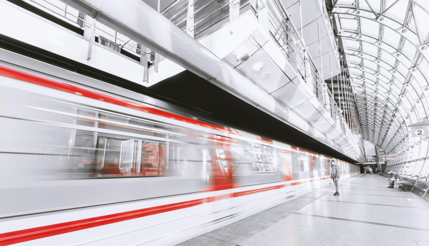 Moderna vilciena stacija ar stikla jumtu. Priekšplānā redzams ātrumā garām braucošs balts vilciens ar sarkanu svītru, fonā – cilvēki gaida uz perona.