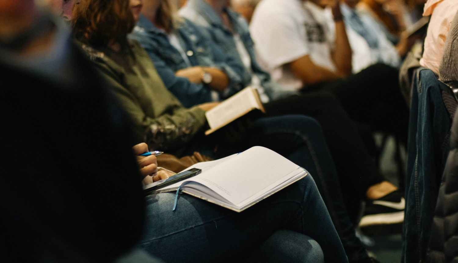 Grupa cilvēku sēž auditorijā vai seminārā, daži raksta piezīmes kladēs, citi lasa grāmatas. Priekšplānā redzams cilvēks ar atvērtu kladi un pildspalvu rokā.