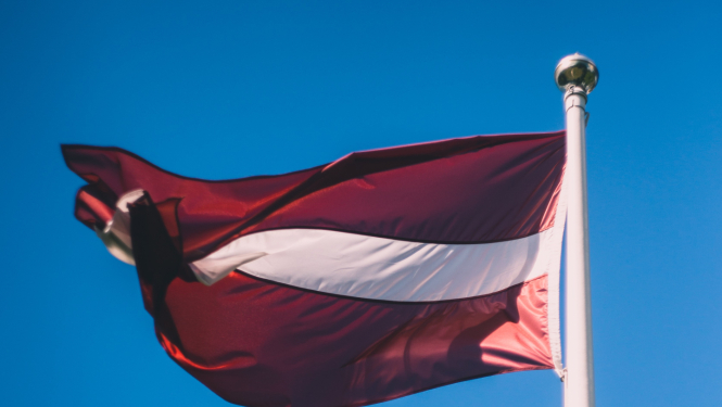 Attēlā redzams Latvijas valsts karogs, kas plīvo vēja karoga mastā, zilas debess fonā.