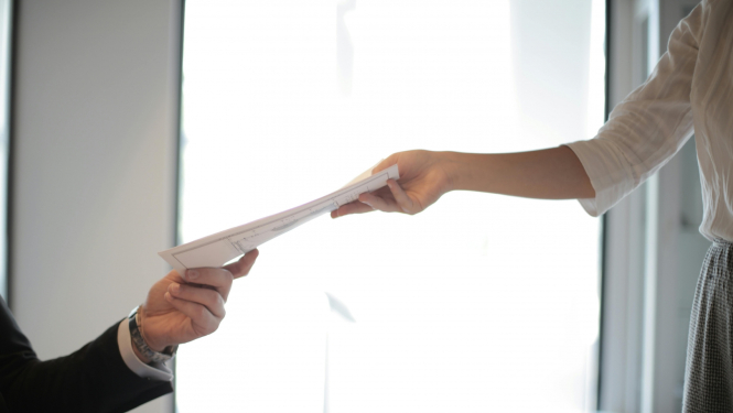 Divi cilvēki pie galda nodod viens otram dokumentus – viens sniedz dokumentu, otrs to pieņem.