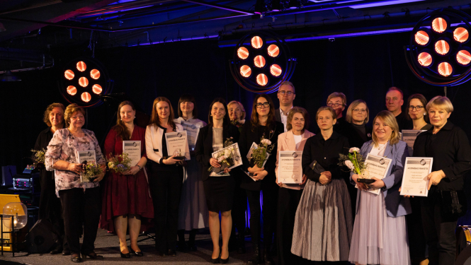 Grupas foto ar cilvēkiem, kas stāv uz skatuves un tur rokās atzinības rakstus un ziedus. Fonā redzami spilgti skatuves prožektori.