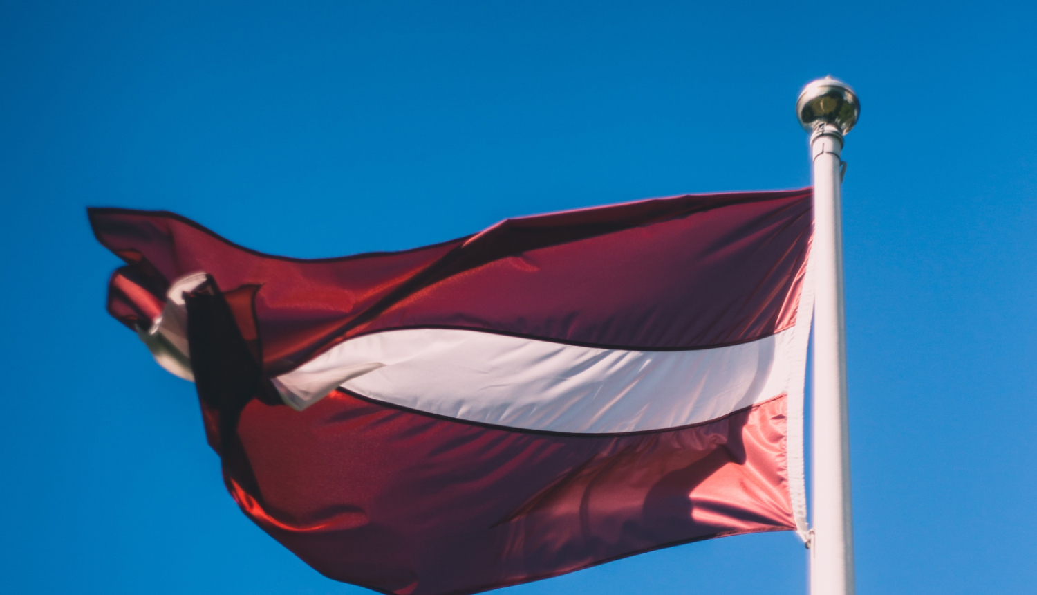 Attēlā redzams Latvijas valsts karogs, kas plīvo vēja karoga mastā, zilas debess fonā.