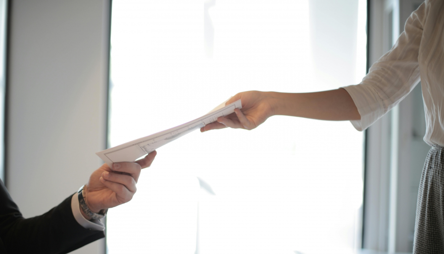 Divi cilvēki pie galda nodod viens otram dokumentus – viens sniedz dokumentu, otrs to pieņem.