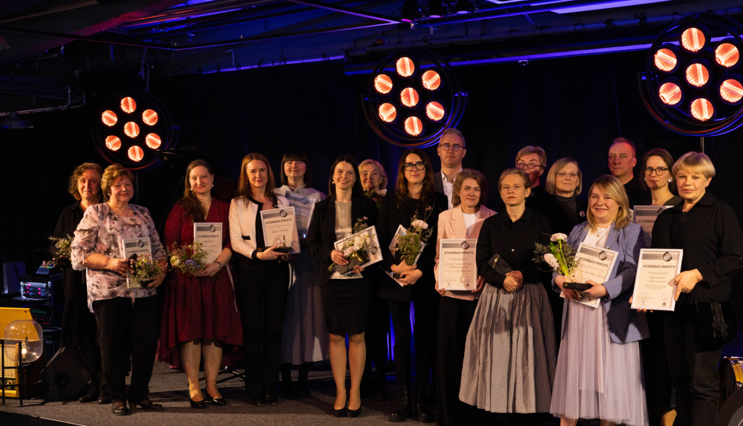 Grupas foto ar cilvēkiem, kas stāv uz skatuves un tur rokās atzinības rakstus un ziedus. Fonā redzami spilgti skatuves prožektori.